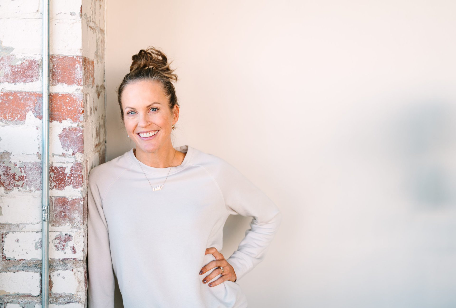 Lydia Fine standing inside a room wearing a cream crewneck sweatshirt leaning against a brick wall with her hand on her hip.